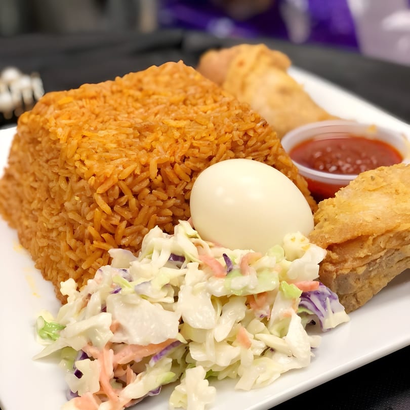 Accra Girls Restaurant's jollof rice, coleslaw, hard-boiled egg, fried fish, and red sauce on a white plate.