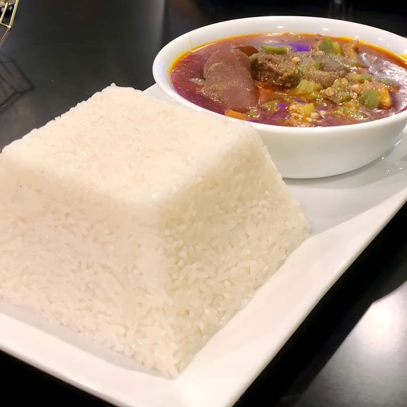 A white bowl of red stew with meat and okra, served with a rectangular mound of white rice.