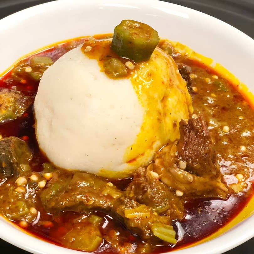 A white bowl of Accra Girls Restaurant's fufu and okra stew with meat, garnished with a piece of okra.